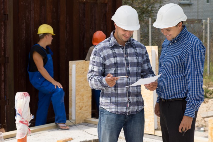 Engineer and architect wearing hardhats standing discussing paperwork on a construction site with builders working behind them